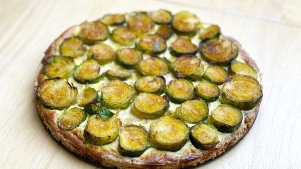 Das Bild zeigt eine runde Tarte mit gerösteten Rosenkohlstücken auf einem Holzuntergrund. Die Tarte hat einen goldbraunen Rand und ist gleichmäßig mit den Kohlscheiben belegt.