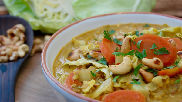 KI generiert: Das Bild zeigt eine Schüssel mit einem Gemüsecurry, das mit Karotten, Kohl, Cashewnüssen und frischen Kräutern garniert ist. Im Hintergrund sind weitere Cashewkerne und ein Stück Kohl zu sehen.